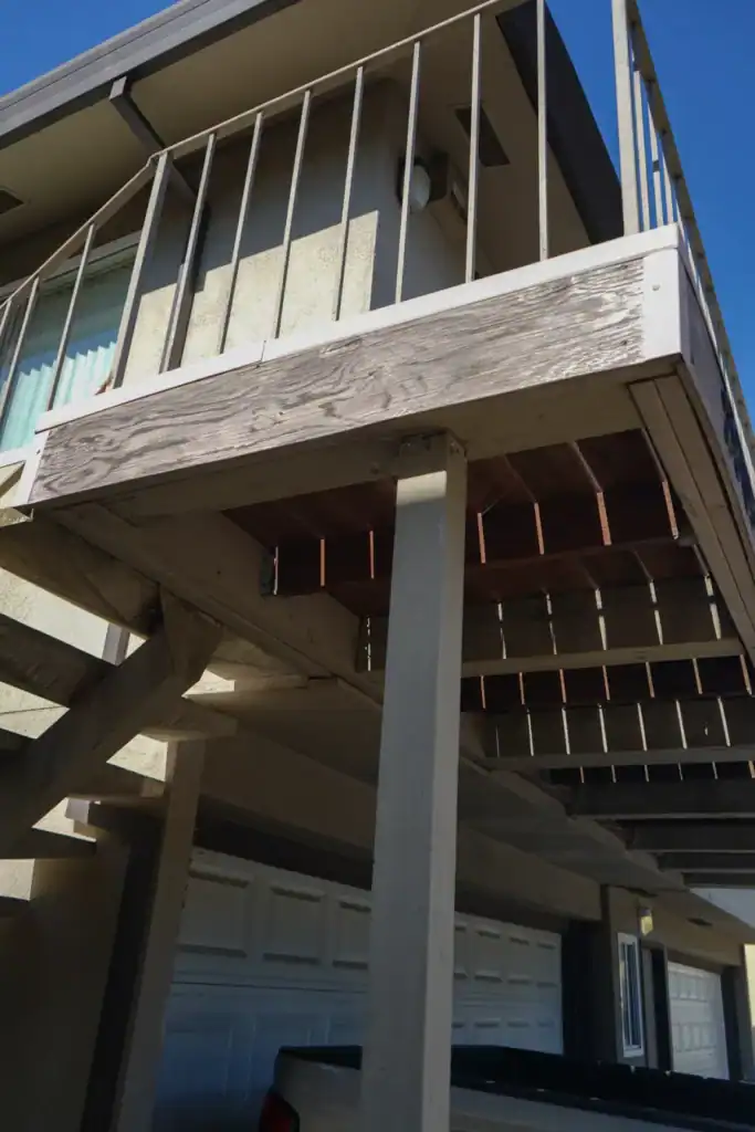 Weathered rim beam and exposed joists under an elevated entry balcony, with daylight showing through the gaps in the decking
