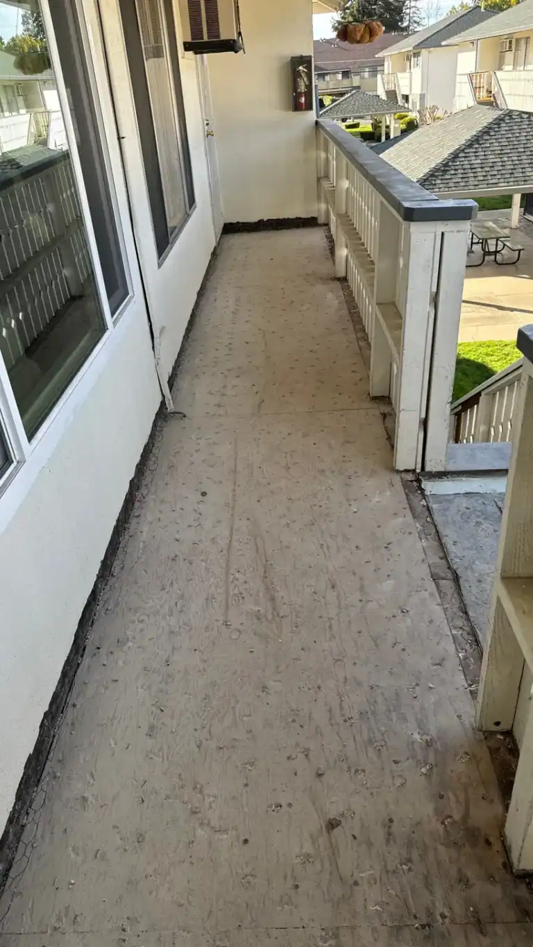 Long view down an apartment balcony stripped to bare sheathing, with the wall line opened up for a new termination