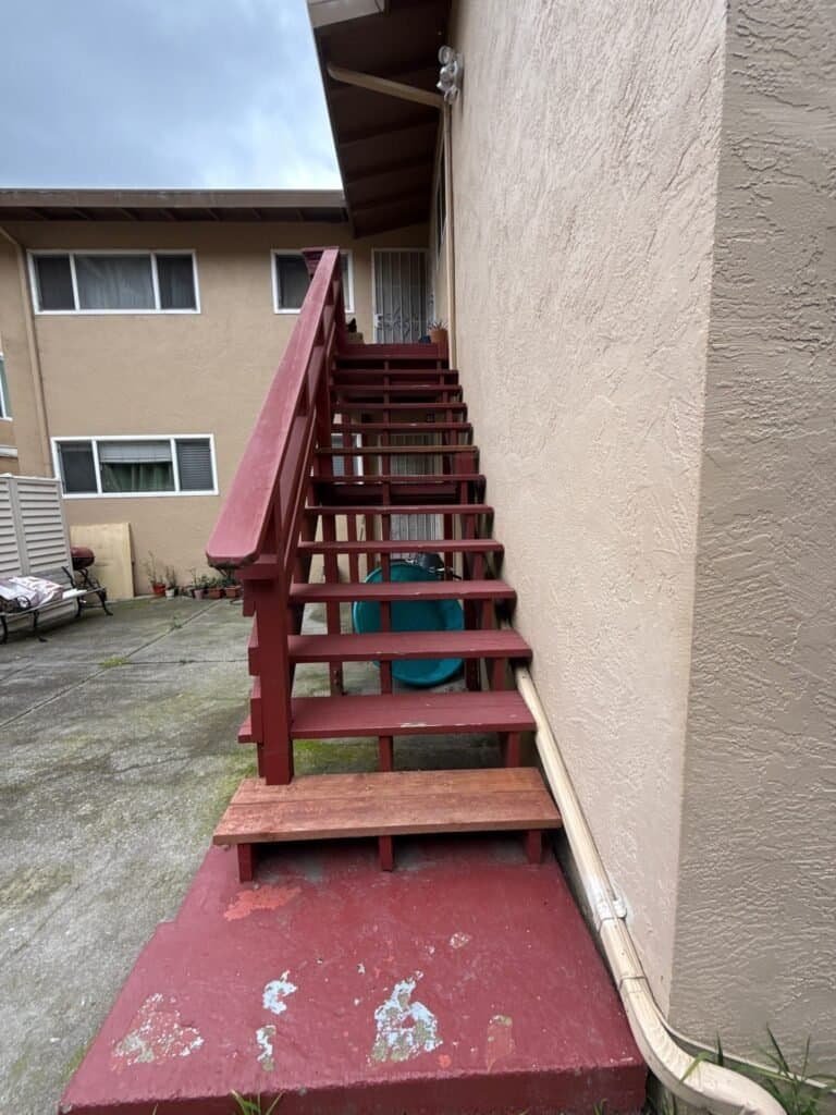 Painted wood apartment stair in Oakland with a flat cap board handrail and a patched bottom tread before replacement