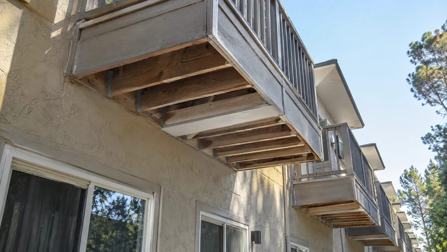 Underside of a wood balcony showing exposed joists and metal hangers on a stucco wall