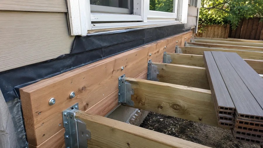New ledger bolted to the wall with flashing membrane above it, joist hangers set and decking stacked
