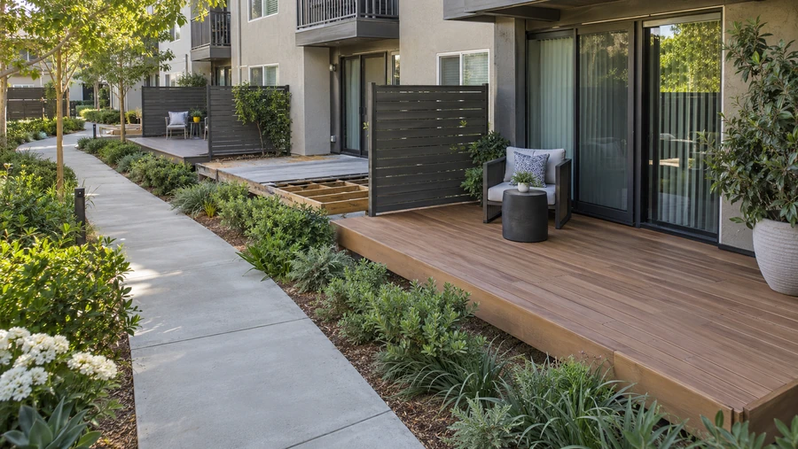 Row of ground floor apartment patio decks along a walkway, one with boards removed to expose joists