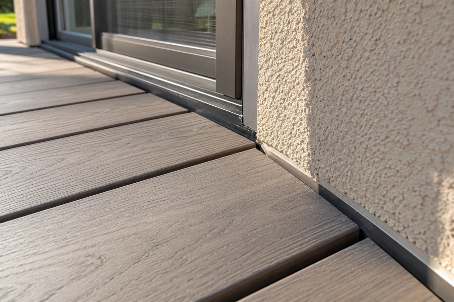 Composite deck boards ending beside a stucco wall and a sliding door, with a small gap between the board ends and the wall